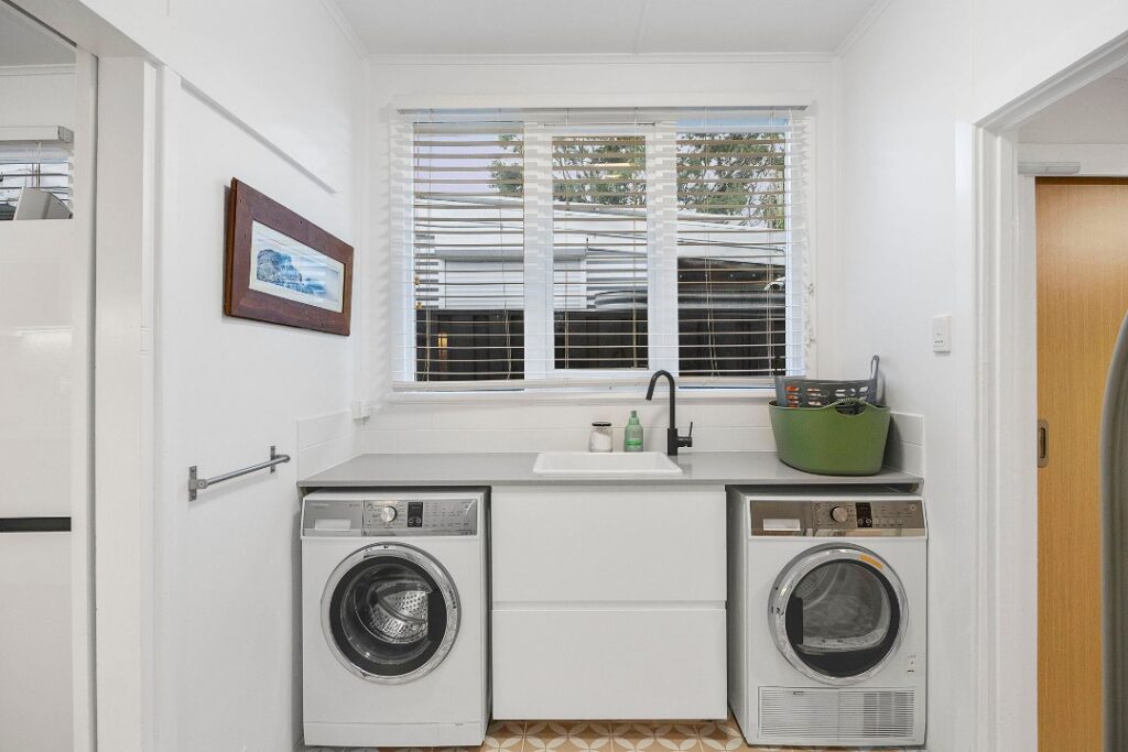 Bright laundry room with a washer, dryer, sink, and large window with white blinds.