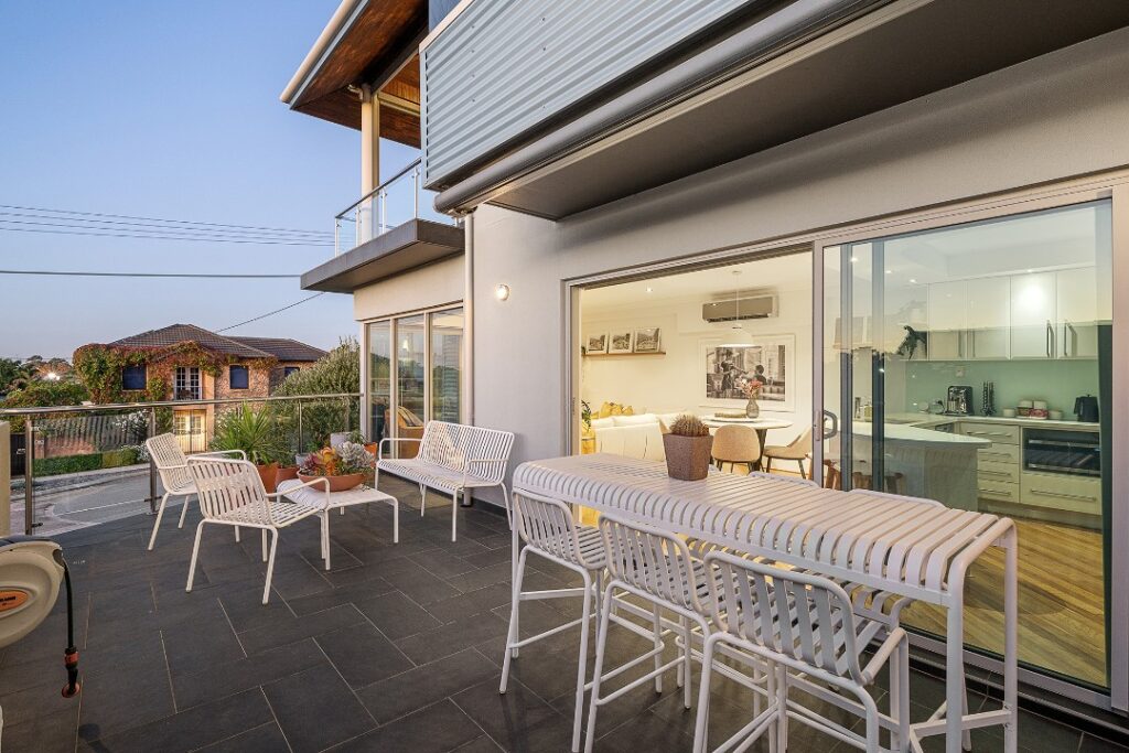 Spacious balcony with white outdoor seating, a bar table, and open views into a modern apartment.