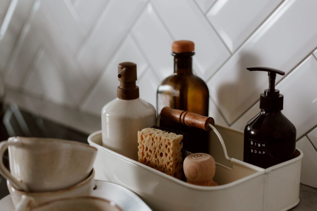 Close-up of a white caddy holding kitchen cleaning supplies, including wooden-topped soap dispensers, a natural sponge with scrub brush, and a dark glass "Dish Soap" pump bottle, against white herringbone tile backsplash