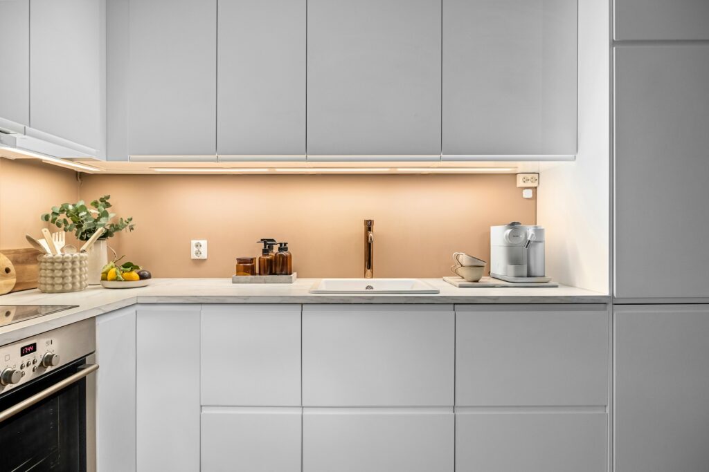 Modern grey kitchen with marble-look countertop, warm under-cabinet lighting, coffee machine, ceramic soap bottles, chopping board with fresh produce, and stainless steel oven with digital display