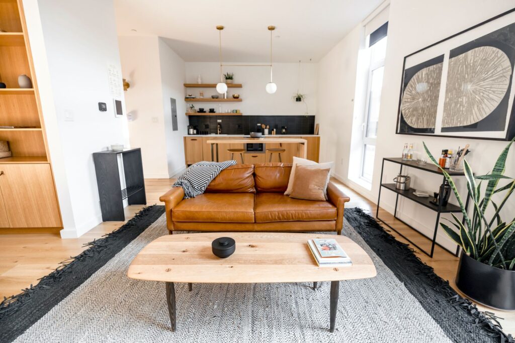 Modern minimalist living room with a tan leather sofa styled with a black and white striped throw and cushions, a light wood coffee table in front, black and white geometric rug, open-plan kitchen with pendant lighting behind, and an abstract artwork on the wall to the right