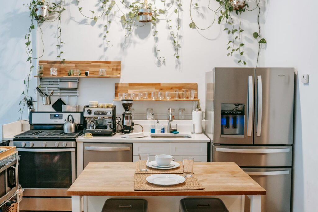 Industrial style kitchen with stainless steel fridge and appliances, espresso machine and grinder on white countertop, open wood shelving with glassware, trailing plants hanging above, and wooden dining table set with a single place setting