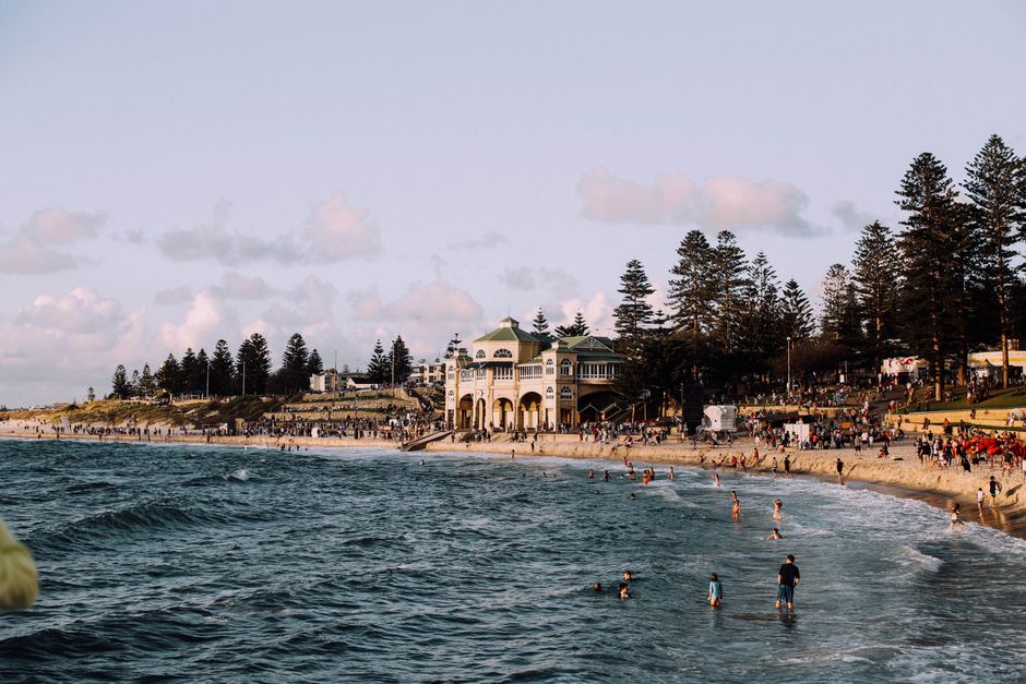 Busy WA coastal beach with swimmers and pine trees, holiday destination appeal.