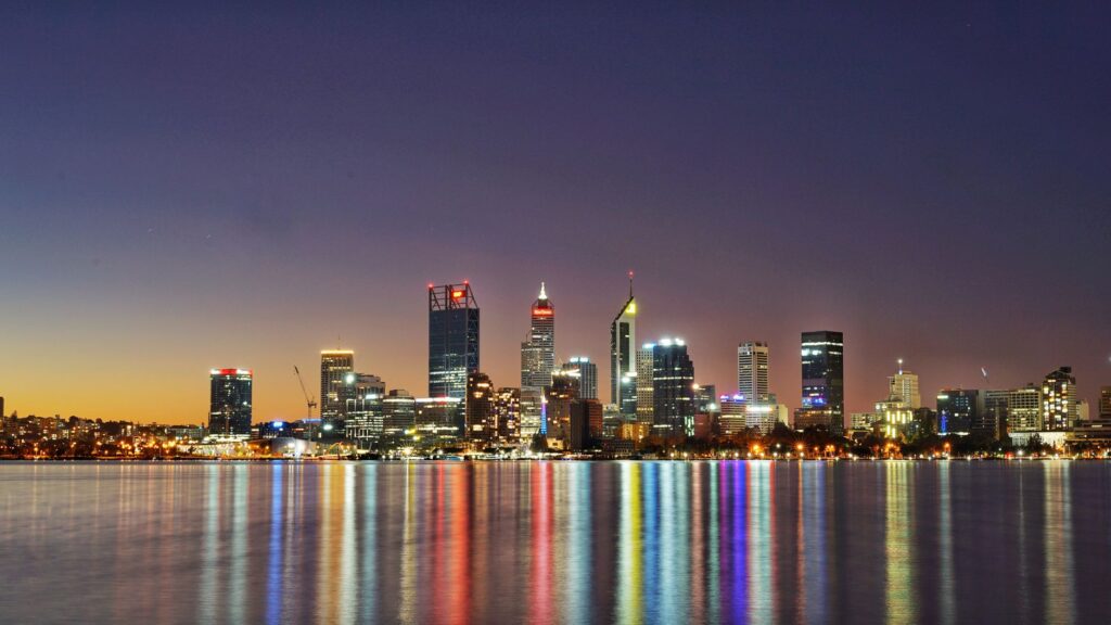 Perth city skyline at twilight reflecting in calm water with coloured lights.