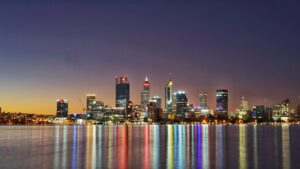 Perth city skyline at twilight reflecting in calm water with coloured lights.