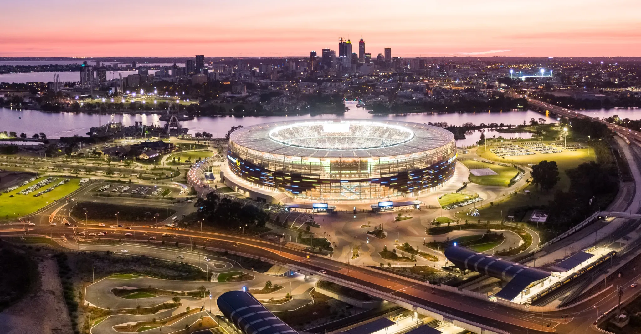 Optus Stadium illuminated at dusk, with the Swan River and Perth skyline behind it